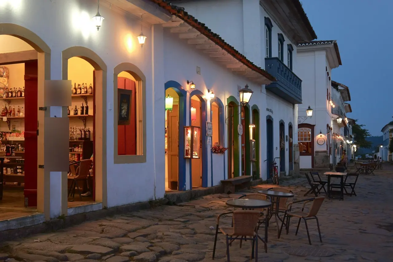 Rua de comércio com lojas iluminadas e mesas na calçada ao entardecer