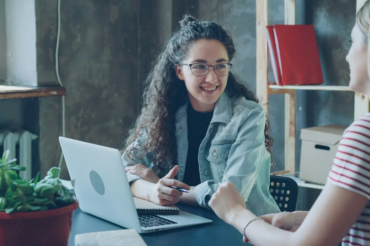 Mulher sorrindo diante do notebook enquanto conversa com outra pessoa