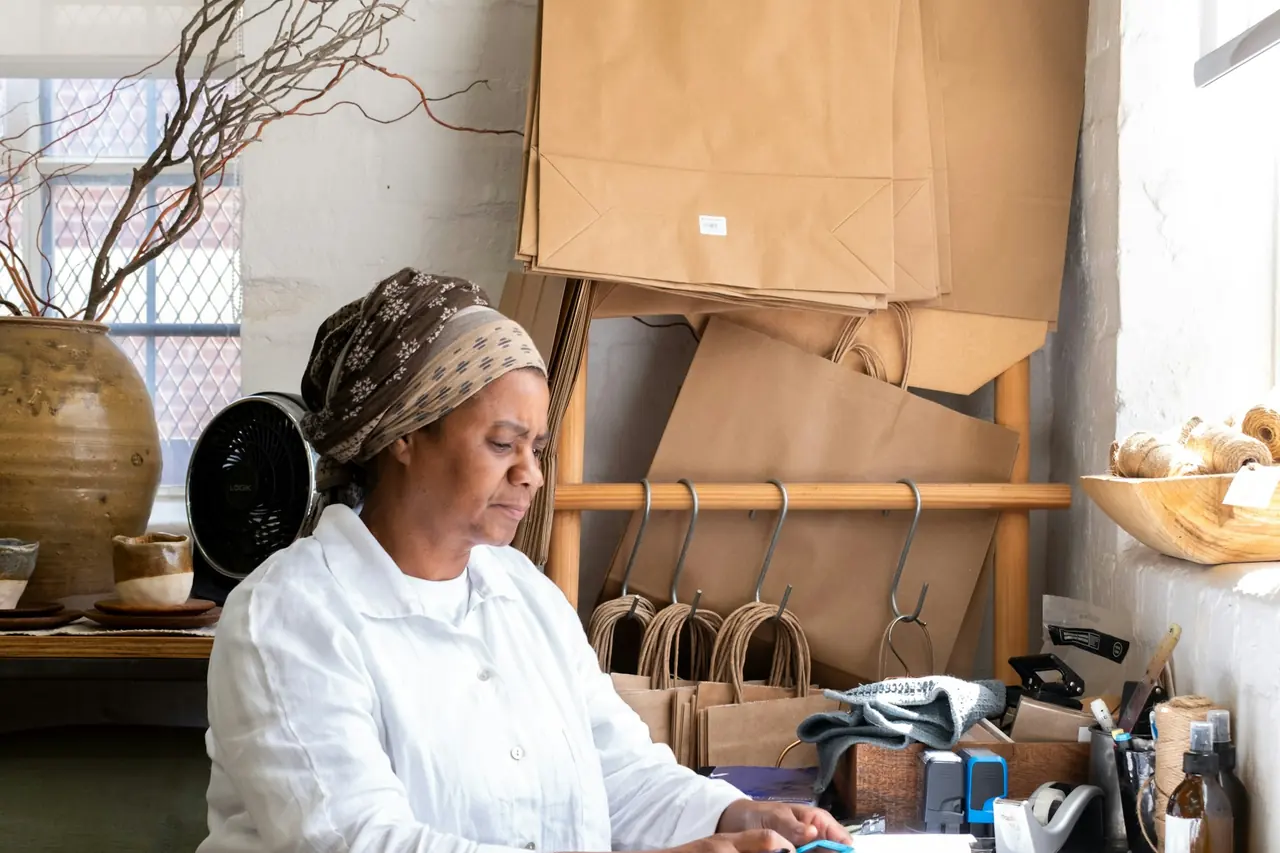 Artesã trabalhando no ateliê entre sacolas de papel e materiais