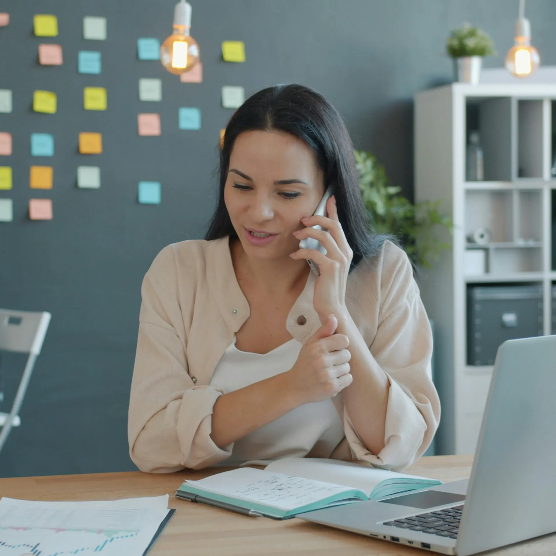 Executiva sorrindo ao telefone na mesa do escritório, com notebook e caderno