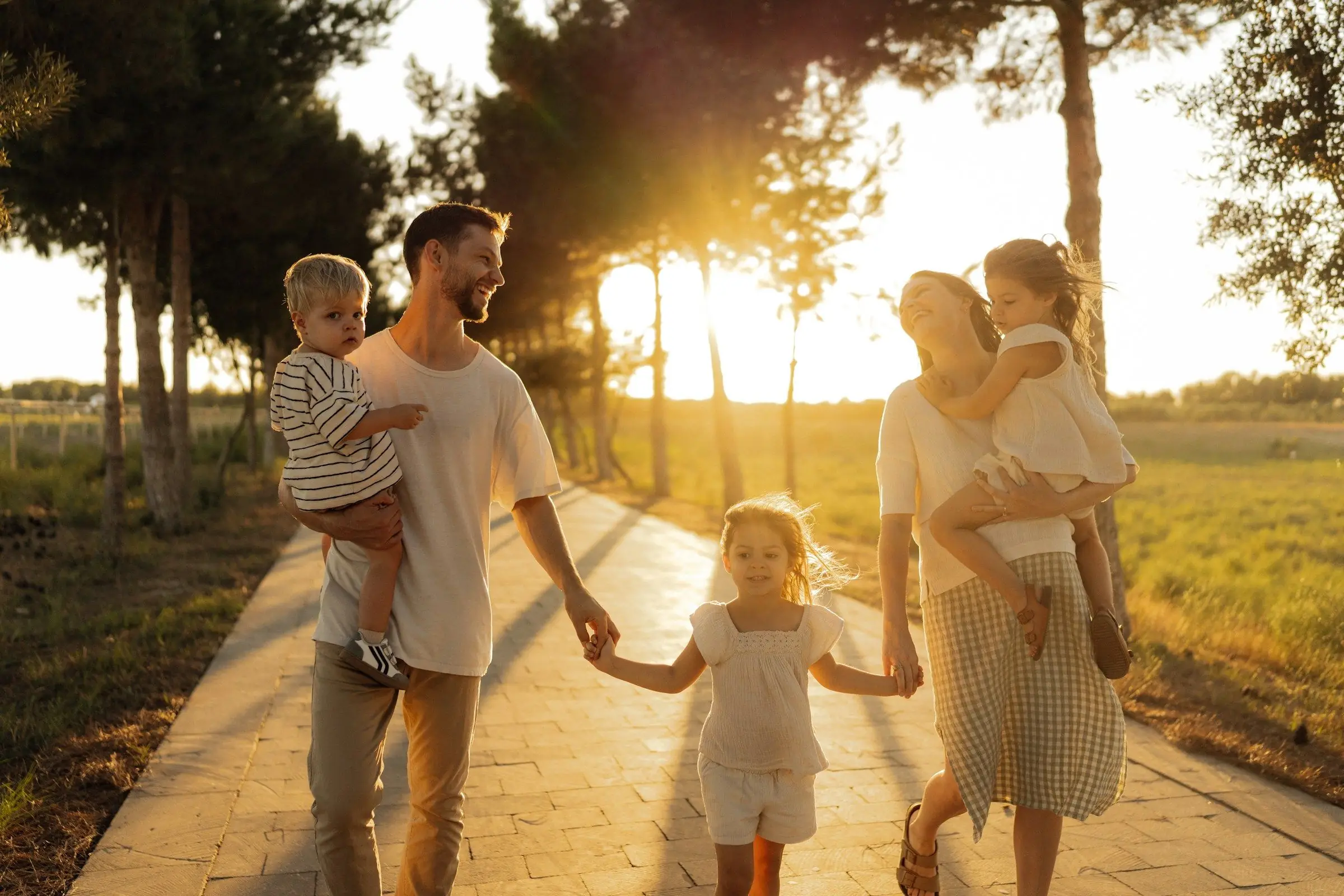 Casal com três crianças caminhando de mãos dadas por uma alameda de árvores ao pôr do sol