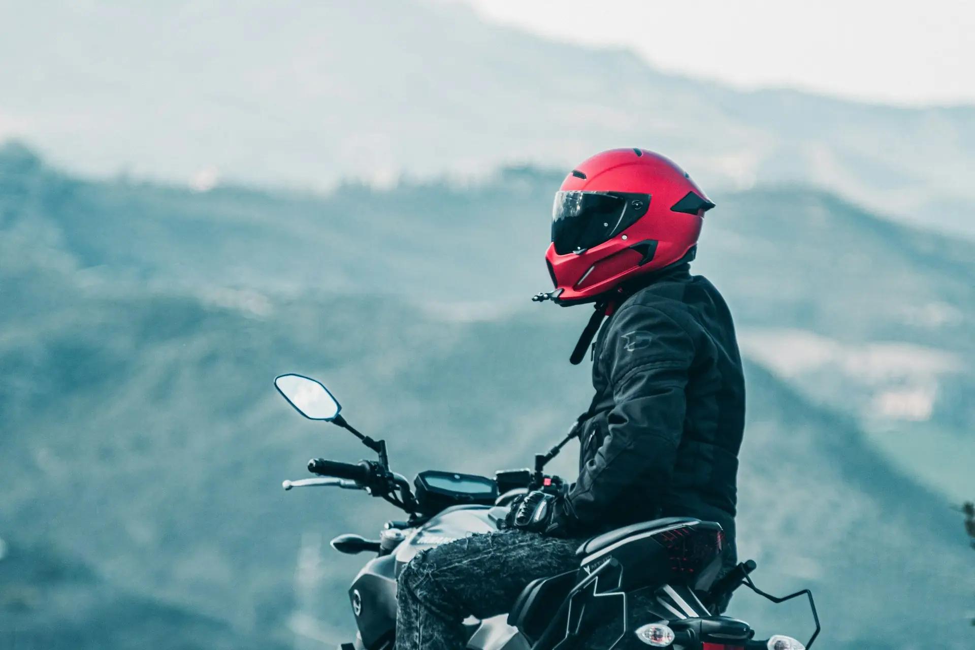 Motociclista de capacete vermelho parado na moto, com montanhas ao fundo