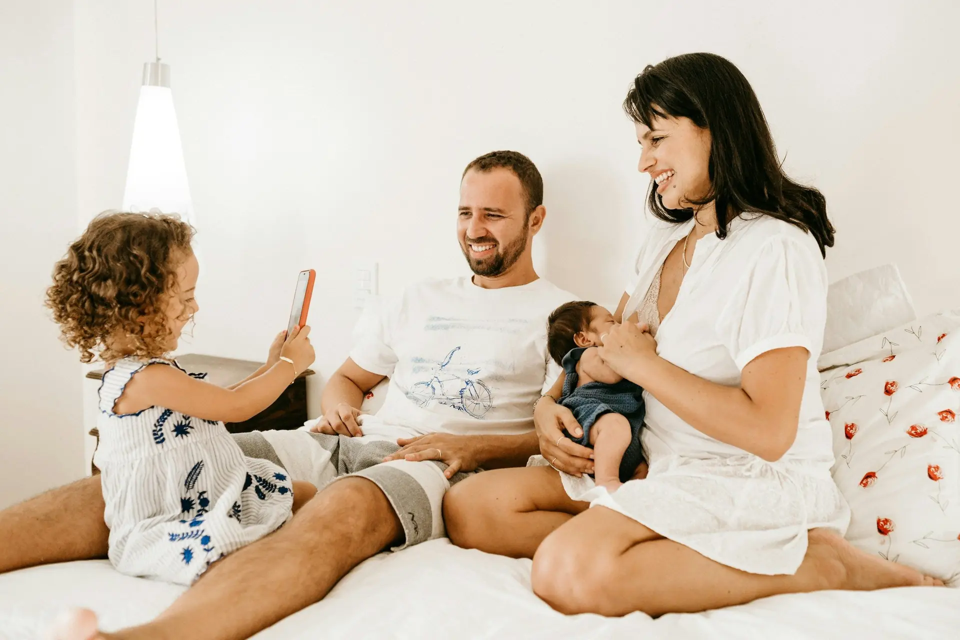 Família reunida na cama de casa: pai, mãe com um bebê no colo e uma menina segurando um celular